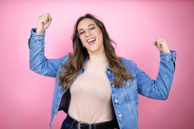 Soyutlanmış pembe arka planda kot ceket giyen genç ve güzel bir kadın mutlu ve heyecanlı. Kollarını kaldırmış, gülümseyen ve başarı için çığlık atan kazanan bir jest yapıyor..