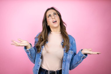 Soyutlanmış pembe arka planda kot ceket giyen genç ve güzel bir kadın. Kolları ve elleri havada, kafası karışmış bir ifade.