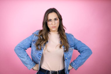 İzole edilmiş pembe arka plan üzerinde kot ceket giyen genç ve güzel bir kadın şüpheci ve gergin, yüzünde tasvip etmeyen bir ifade belimde kollarıyla
