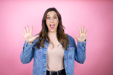 İzole edilmiş pembe arka planda kot ceket giyen genç ve güzel bir kadın 10 numaralı parmaklarıyla işaret ederken kendinden emin ve mutlu bir şekilde gülümsüyor.