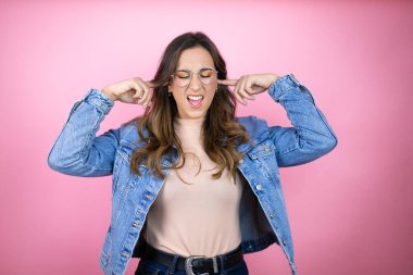 İzole edilmiş pembe arka planda kot ceket giyen genç ve güzel bir kadın yüksek sesli müzik sesi için parmaklarıyla kulaklarını kapatıyor. Sağır kavramı.