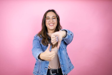 Soyutlanmış pembe arka planda kot ceket giyen genç ve güzel bir kadın baş parmaklarını yukarı aşağı hareket ettiriyor, anlaşmazlık ve anlaşma ifadesi takınıyor. Çılgın çatışma