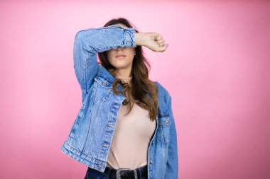 İzole edilmiş pembe arka planda kot ceket giyen genç ve güzel bir kadın. Gözleri kollarıyla kaplayarak neşeli ve komik gülümsüyor. Kör kavram.