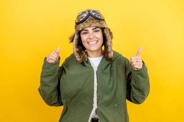 Sarı arka plan başarısı tabelasının üzerine gözlük ve ceket takmış, el hareketleriyle olumlu jest yapan, gülümseyen ve mutlu, klasik pilot kaskı takan genç güzel bir havacı kadın. Neşeli ifade ve kazanan jest.