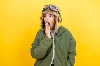 Sarı arka plan üzerinde gözlük ve ceket takan, ağzı elle kaplayan, şok olmuş ve hata yapmaktan korkan, klasik pilot kaskı takan genç, güzel bir havacı kadın. şaşırmış ifade