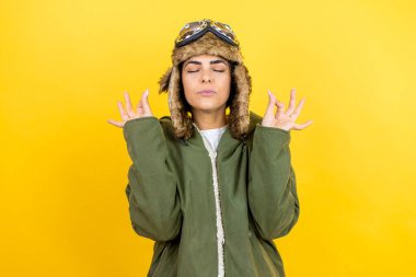 Sarı arka planda gözlüklü ve ceketli, klasik pilot kaskı takan genç, güzel bir havacı kadın. Gözleri kapalı, meditasyon yaparken parmaklarıyla gülümsüyor.