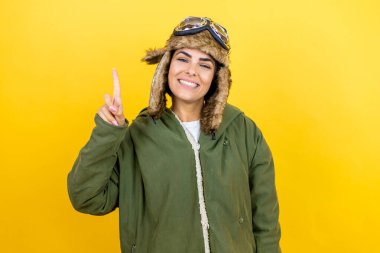 Sarı arka planda gözlüklü, klasik pilot kaskı takan genç ve güzel bir havacı kadın bir numarayı gösterirken kendinden emin ve mutlu bir şekilde gülümsüyor.