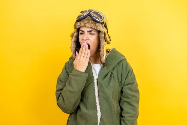 Sarı zemin üzerinde gözlük ve ceket olan klasik pilot kaskı takan genç, güzel bir havacı kadın esnemekten sıkılmış, ağzı eliyle kapatmaktan sıkılmış. Huzursuz ve uykulu..