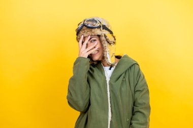 Sarı arka plan üzerinde gözlük ve ceketi olan klasik pilot kaskı takan genç, güzel bir havacı kadın. Şok olmuş bir şekilde yüzünü ve gözlerini kapayarak utanmış bir ifadeyle parmaklarının arasından bakıyor.