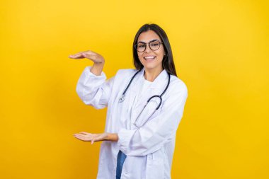 Steteskop takan genç esmer kadın izole edilmiş sarı arka planda duruyor. Ellerinde büyük ve büyük boy işareti var. Ölçüm sembolü. Gülümsüyor. Ölçüm konsepti.