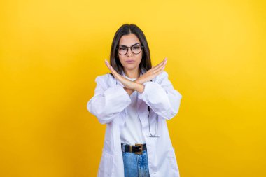 Young brunette doctor woman wearing stethoscope standing over isolated yellow background Rejection expression crossing arms doing negative sign, angry face clipart