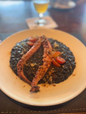 Kirazlı domateslerle süslenmiş zengin, kremalı mürekkep risottosunun üzerine zarif bir şekilde yerleştirilmiş ahtapot dokunaçlarından oluşan bir gurme tabağı, bulanık bir içecek zeminiyle beyaz bir tabakta servis ediliyor..