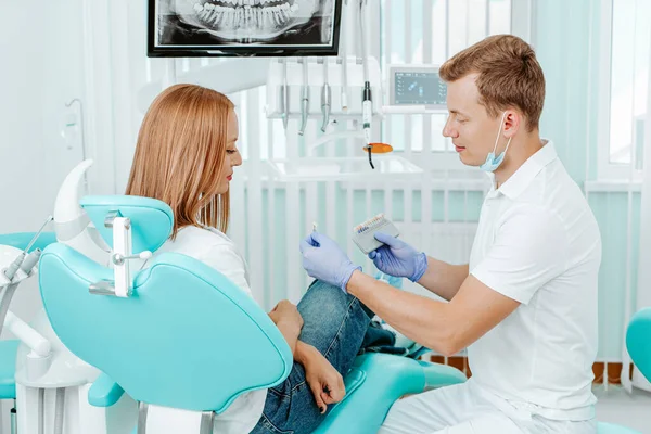 stock image Doctor dentist chooses the whitening color for teeth the young attractive patient in stomotology clinic. Hands dentist with teeth color palette next by face. Smile healthy teeth concept