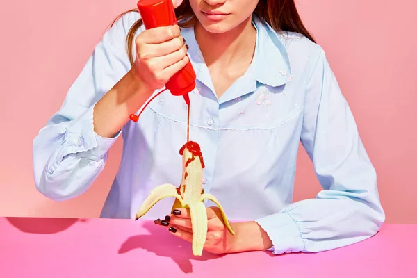 增加味道 食品流行艺术摄影 年轻女子品尝香蕉与番茄酱分离的粉红色背景 复古风格的内饰 — 图库照片