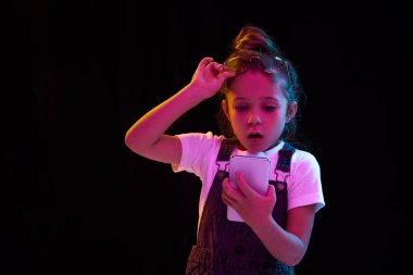 Şaşırmış ilkokul çağındaki kız, koyu arkaplanda izole bir şekilde telefon ekranına bakan öğrenci. Çocuk duyguları, moda, yüz ifadeleri kavramı. İlan için alanı kopyala