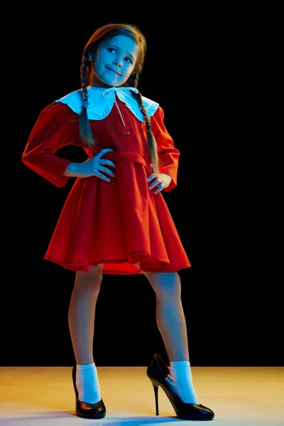 ファッションショー 赤いお祝いのドレスと暗い背景に隔離されたかかとを身に着けているピグテールと美しいかわいい女の子の肖像画 子供の感情 子供の心理学の概念 — ストック写真