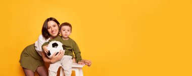 Parlak sarı arka planda izole edilmiş futbol topu tutan sevimli oğluyla gülümseyen genç anne. Aile, sevgi, annelik ve anneler günü konsepti. İlan için kopyalama alanı olan broşür