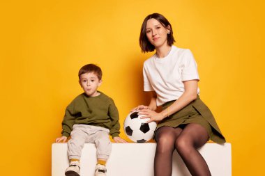 Çocuklu çekici bir genç kadın, annesi ve futbol topu tutan küçük oğlu parlak sarı arka planda izole edilmiş. Aile, sevgi, annelik ve anneler günü konsepti. İlan için alanı kopyala