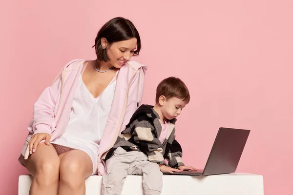 Watching cartoon. Beautiful young woman and little boy, mom and son using laptop isolated on pink background. Mothers Day celebration. Concept of family, childhood, motherhood, sincere emotions