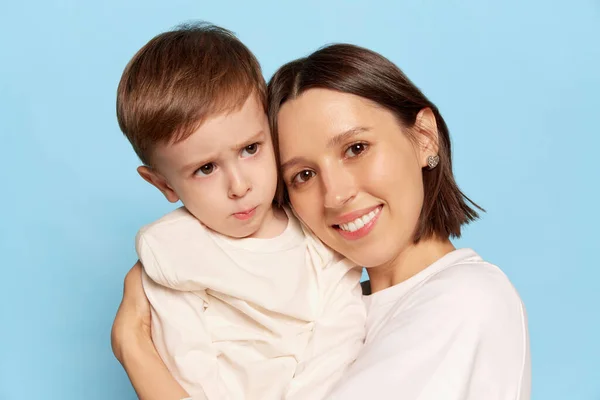 Tenderness. Half-length portrait of beautiful happy woman hugging her little son, kid isolated on blue background. Family, love, motherhood and Mothers day concept.
