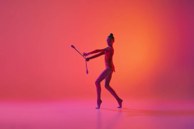 Genç ve çekici bir kızın stüdyo çekimi, neon ışık filtresiyle izole edilmiş spor ekipmanlarıyla ritmik jimnastik eğitimi. Dans, müzik, duygular, zorluklar, motivasyon ve reklam