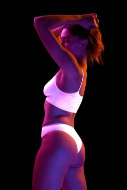 Beyaz iç çamaşırlarıyla poz veren ince vücutlu güzel bir kadının portresi neon ışıkta siyah arka planda izole edilmiş. Güzellik, vücut ve cilt bakımı, sağlık, plastik cerrahi, kozmetik, spor