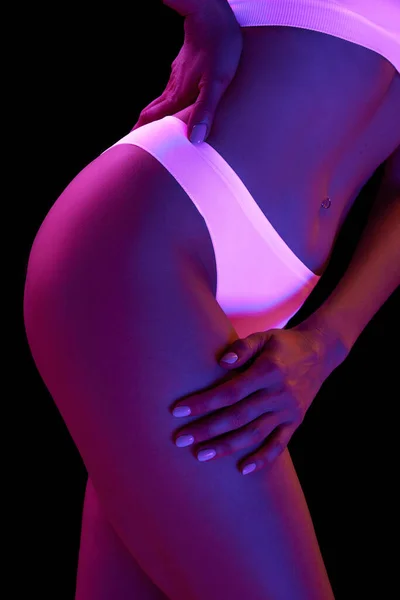 Cropped image of slim female body in white underwear over black background in neon light. Buttocks and smooth legs. Concept of beauty, body and skin care, health, plastic surgery, cosmetics