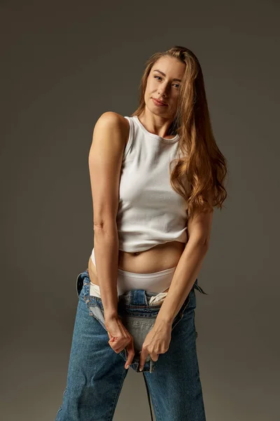 Portrait of beautiful woman posing in casual clothes, jeans and white top over dark grey studio background. Comfort. Concept of beauty, fashion, style, body and skin care, health, cosmetics