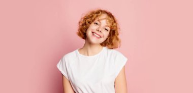 Kızıl saçlı genç kızın yarım boy portresi pembe stüdyo arka planında duygularını ifade eden öğrenci. Reklam için boşluk kopyala. Mutlu ve sakin görünüyor..