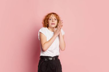 Kıvırcık saçlı, beyaz tişörtlü, gözleri, pembe stüdyo arka planında izole edilmiş kameraya bakan korkmuş genç kız. İnsani duygular, yüz ifadesi, güzellik konsepti. İlan için alanı kopyala