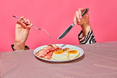 Female hands with fork and knife eating English breakfast with fried eggs and bacon . Vintage, retro style interior. Food pop art photography. Complementary colors. Copy space for ad, text clipart