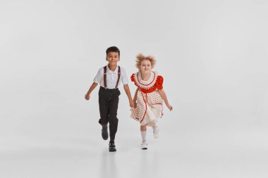 Güzel küçük çocukların portresi, klasik kıyafetlerle koşan kız ve erkek, gri arka planda izole bir şekilde birlikte oynuyorlar. Çocukluk kavramı, yaratıcılık, eski moda, arkadaşlık, sanat