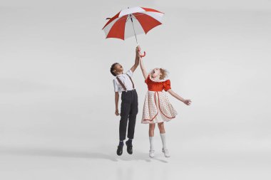 Gri arka planda izole edilmiş şemsiyeyle atlayan klasik elbiseli kız ve erkek çocukların portresi. Çocukluk kavramı, yaratıcılık, eski moda, arkadaşlık, sanat