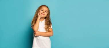 Gülümseyen çocuk. Şirin küçük mutlu kız, uzun kıvırcık saçlı çocuk mavi arka planda izole edilmiş kameraya bakıyor. Çocukların olumlu duyguları, güzelliği, yüz ifadeleri kavramı. İlan için alanı kopyala
