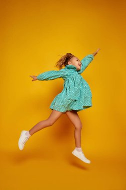 Eğlenmene bak. Mutlu gülümseyen küçük kız, renkli elbiseli çocuk sarı stüdyo arka planında yüksek izole atlama. Çocuklar moda, duygular, tasasız çocukluk, zorluklar, eğitim ve özgürlük kavramı