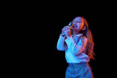 Mikrofonla şarkı söylemek. Duygusal sevimli küçük kız, karanlık stüdyo arka planında neon ışıkta izole edilmiş bir çocuk. Çocuk duyguları, moda, güzellik, okul ve reklam kavramı. Boşluğu kopyala