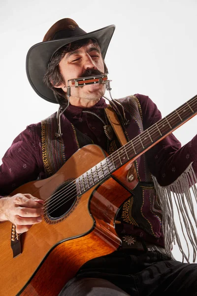 Man with moustaches in country style clothes playing guitar and harmonica, performing isolated over white background. Cowboy style. Concept of music, creativity, inspiration, hobby, lifestyle