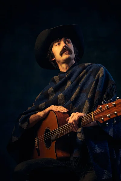 Portrait of man with moustaches in vintage clothes and black hat posing with guitar isolated over dark blue background. Thoughtful look. Concept of music, creativity, inspiration, hobby, lifestyle