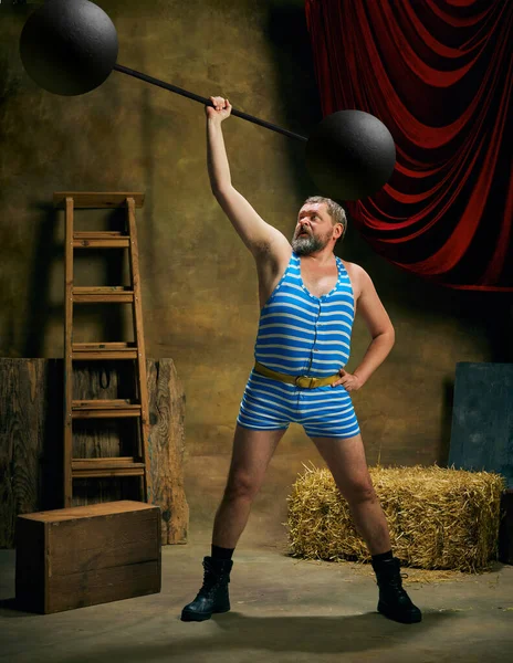Strength. Overweight man, retro circus strongman wearing striped sports swimsuit with barbell over dark circus backstage background. Concept of creative art, fashion, style and inspiration