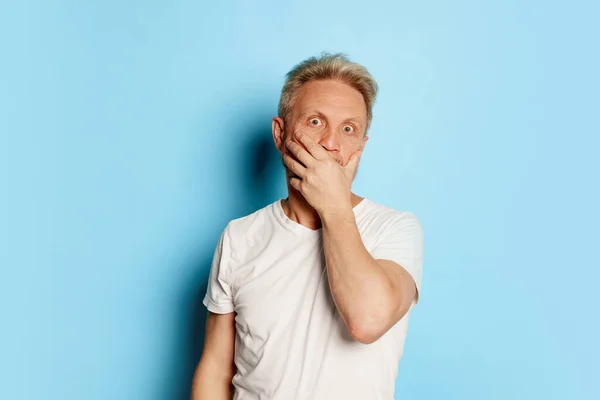 stock image Portrait of mature man in white T-shirt posing, covering mouth in shock isolated over blue studio background. Concept of emotions, facial expression, lifestyle, fashion. Copy space for ad