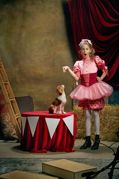 Circus show with animal handler. Little cute beautiful girl in festive dress training funny doggy at vintage circus. Concept of holidays, dreams, art, fashion, vintage style