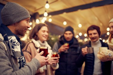 Dostça bir parti. Emotif gülümseyen erkekler ve kadınlar ellerinde kupalar dolusu sıcak şarapla eğleniyorlar, kış fuarında birlikte vakit geçiriyorlar. Keyif alıyorlar, sıcak giyiniyorlar.