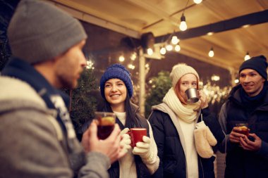 Noel havası. Sıcak giysiler içinde neşeli erkek ve kadınlar kış festivalinde sıcak içeceklerle birlikte vakit geçiriyorlar. Eğleniyorlar, sıcak giyiniyorlar, birbirlerine bakıyorlar ve gülüyorlar.