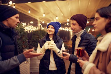 Noel havası. Sıcak giysiler içinde neşeli erkek ve kadınlar kış festivalinde sıcak içeceklerle birlikte vakit geçiriyorlar. Eğleniyorlar, sıcak giyiniyorlar, birbirlerine bakıyorlar ve gülüyorlar.