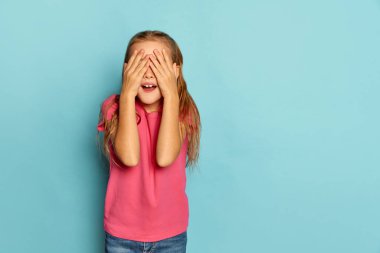 Küçük güzel bir kızın portresi, pembe tişörtlü bir çocuk mavi stüdyo arka planında gözleri kapalı poz veriyor. Saklambaç. Çocukluk kavramı, duygular, yüz ifadesi, yaşam tarzı