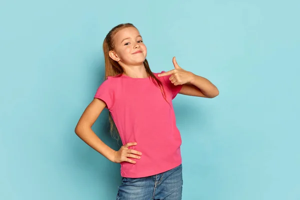 stock image Portrait of little beautiful girl, child in pink T-shirt posing isolated over blue studio background. Proudly pointing at herself. Concept of childhood, emotions, facial expression, lifestyle