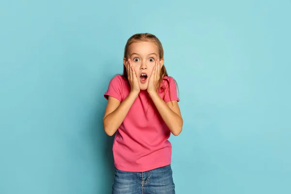 一个漂亮的小女孩的画像 穿着粉色T恤的孩子 脸上带着震惊的表情 与蓝色的工作室背景隔离在一起 童年的概念 面部表情 生活方式 — 图库照片