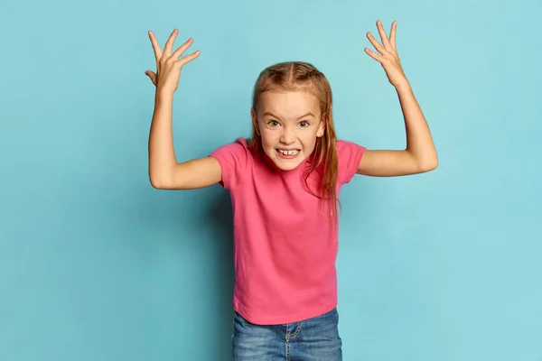 stock image Portrait of little beautiful girl, child in pink T-shirt emotionally posing isolated over blue studio background. Aggressive. Concept of childhood, emotions, facial expression, lifestyle
