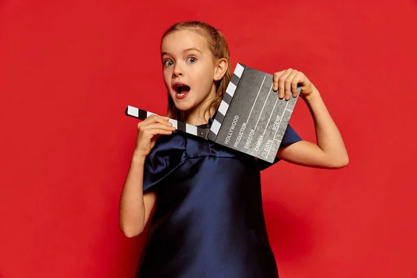 一个漂亮的小女孩的肖像 穿着衣服摆着电影剪贴板的孩子被隔离在红色的工作室背景之外 兴奋极了童年 面部表情 生活方式的概念 — 图库照片