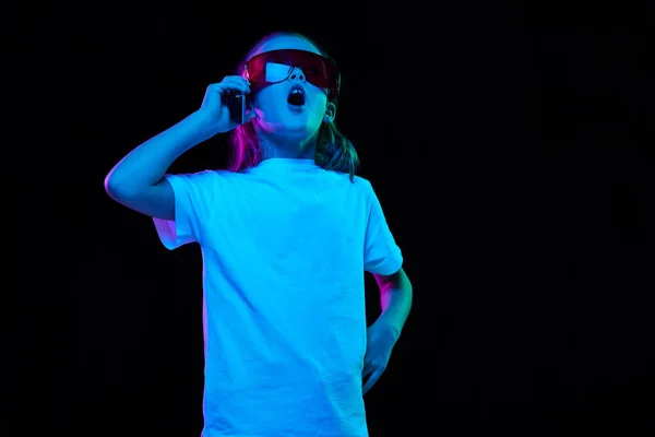 小さな美しい少女の肖像画 白いTシャツの子供の眼鏡や携帯電話のネオンの光の中で黒のスタジオの背景に隔離されたポーズ 子供時代 ライフスタイルの概念 — ストック写真